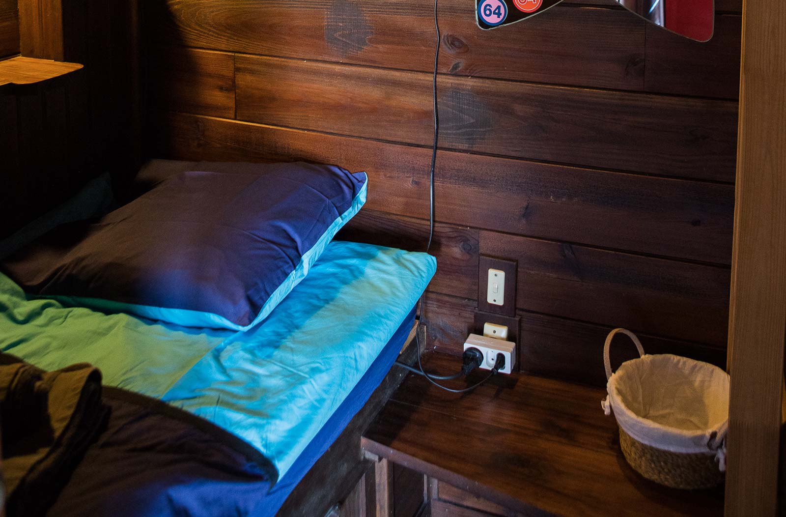 Photo d'une tête de lit avec oreiller et draps marron et bleu. A côté du lit il y a un interrupteur pour la lumière et des prises électriques, mes murs et les meubles sont en bois