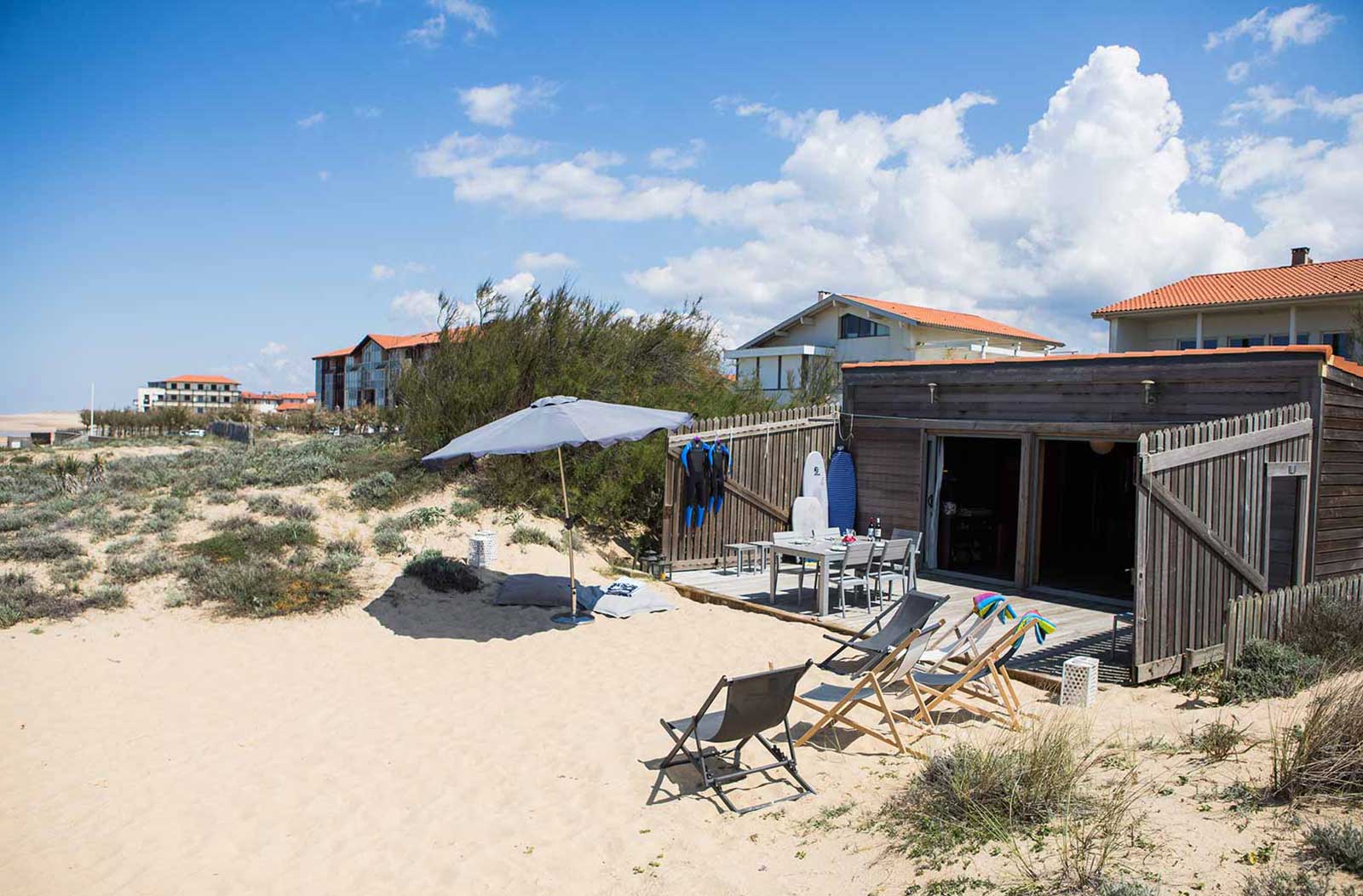 Photo de la façade du cabanon en bois, avec terrasse, table et divers transats posés sur le sable.