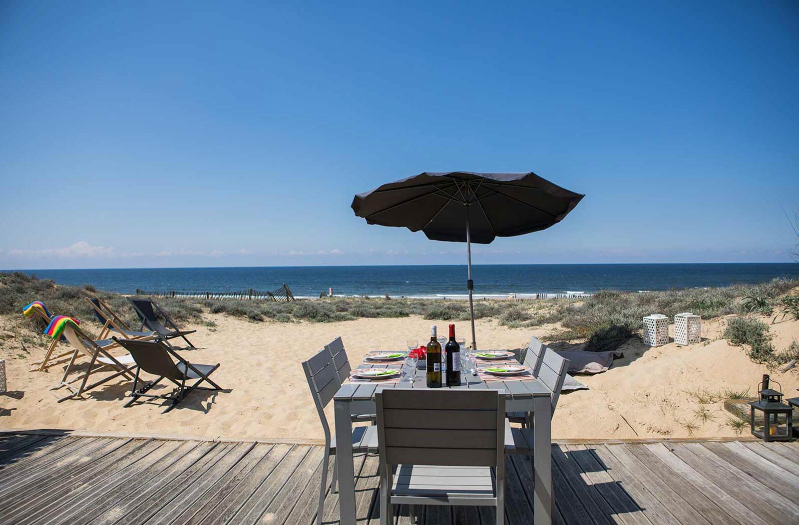 Photo de la terrasse du cabanon, avec vue sur l'océan, table, parasol et transats posés sur le sable.