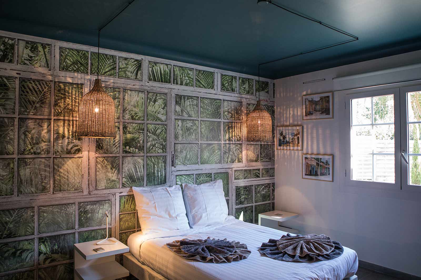 Photo d'une chambre à lit double, le pan de mur de la tête de lit est décoré avec un stickers stylisé jungle, l'autre mur est blanc avec plusieurs cadres photos, et une fenêtre qui donne sur le jardin.