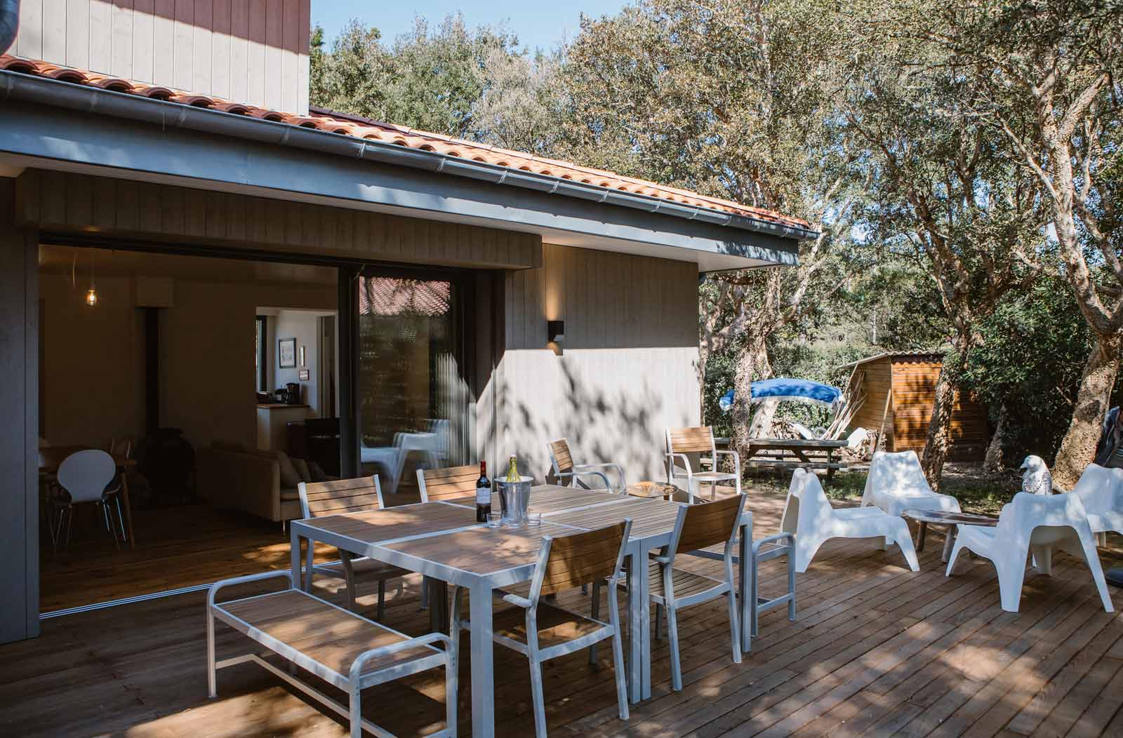 Photo d'une porte fenêtre ouverte de la villa donnant sur la terrasse en bois avec grande table, chaises et tabouret.