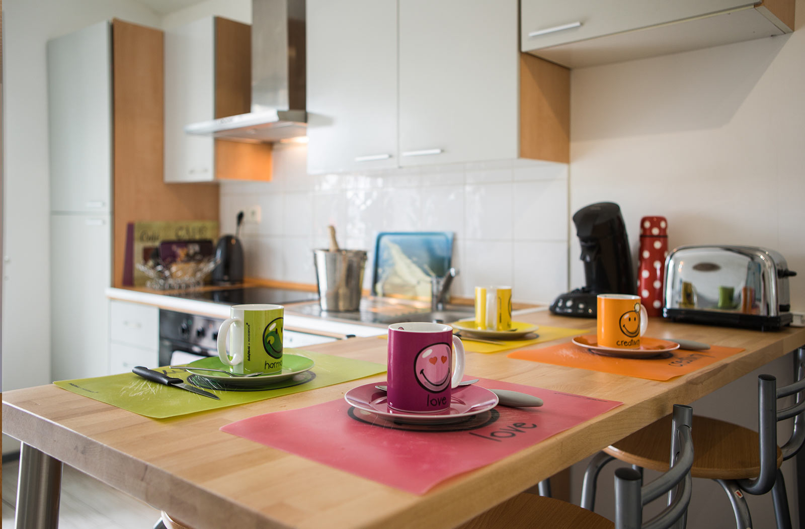 Photo du coin repas de la cuisine, avec sa table en bois, son grille pain et sa machine à café. Le couvert est mise pour le petit déjeuner. En fond on peut apercevoir l'équipement de la cuisine.