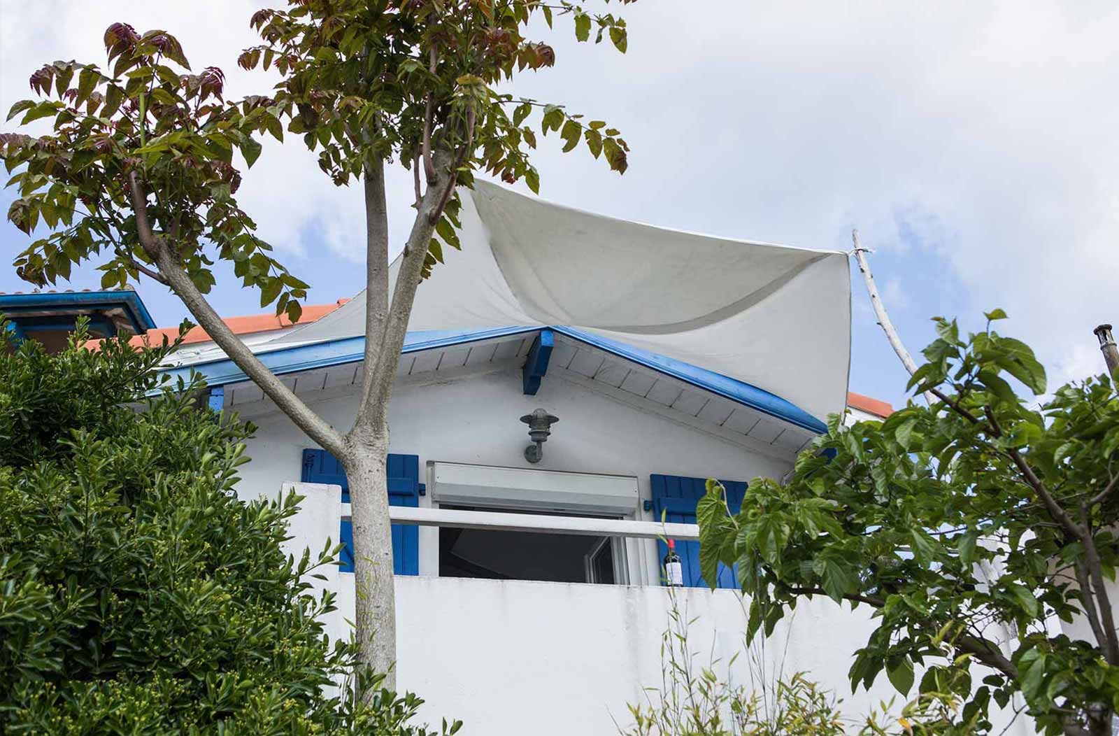 Photo de la façade la maisonnette au milieu des arbres, une fenêtre ouverte donne sur balcon, les volets sont bleus, les murs blancs.