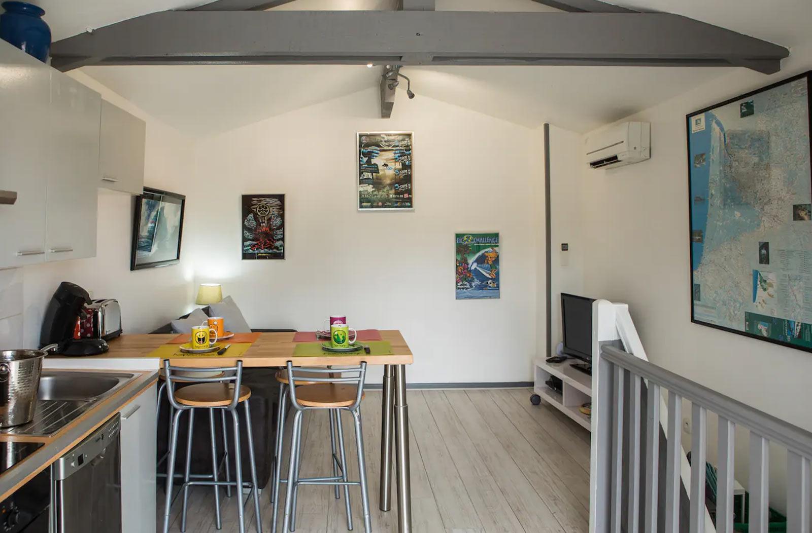 Photo de la cuisine / salon avec table et chaises pour prendre les repas. Le coin cuisine avec plaque à induction, évier, frigo et lave-vaisselle et le coin salon avec banquette et télé posée sur un meuble à roulettes blanc. Les murs de la pièce sont blancs et les poutres apparentes grises.